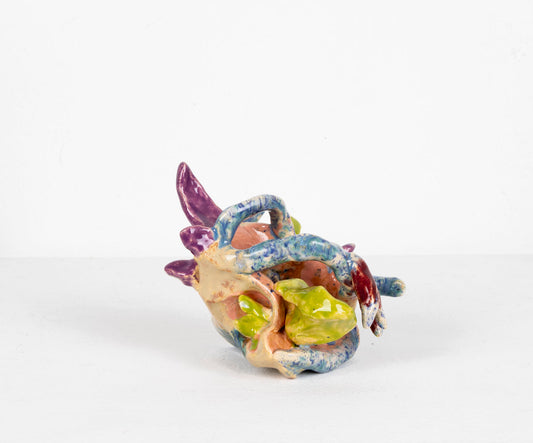 A small abstract ceramic sculpture featuring a cluster of tube and petal shapes crumpled and folded together. From some angles the shapes appear to be contained in or wrapped around a shell. The glaze ranges from deep purple, to pale pink, to a mottled blue flecked with purple. The artist writes: Endless and adrift each oceanic vessel yearns anchor less—beckoned by an impenetrable deep and the promise of a lighthouse.