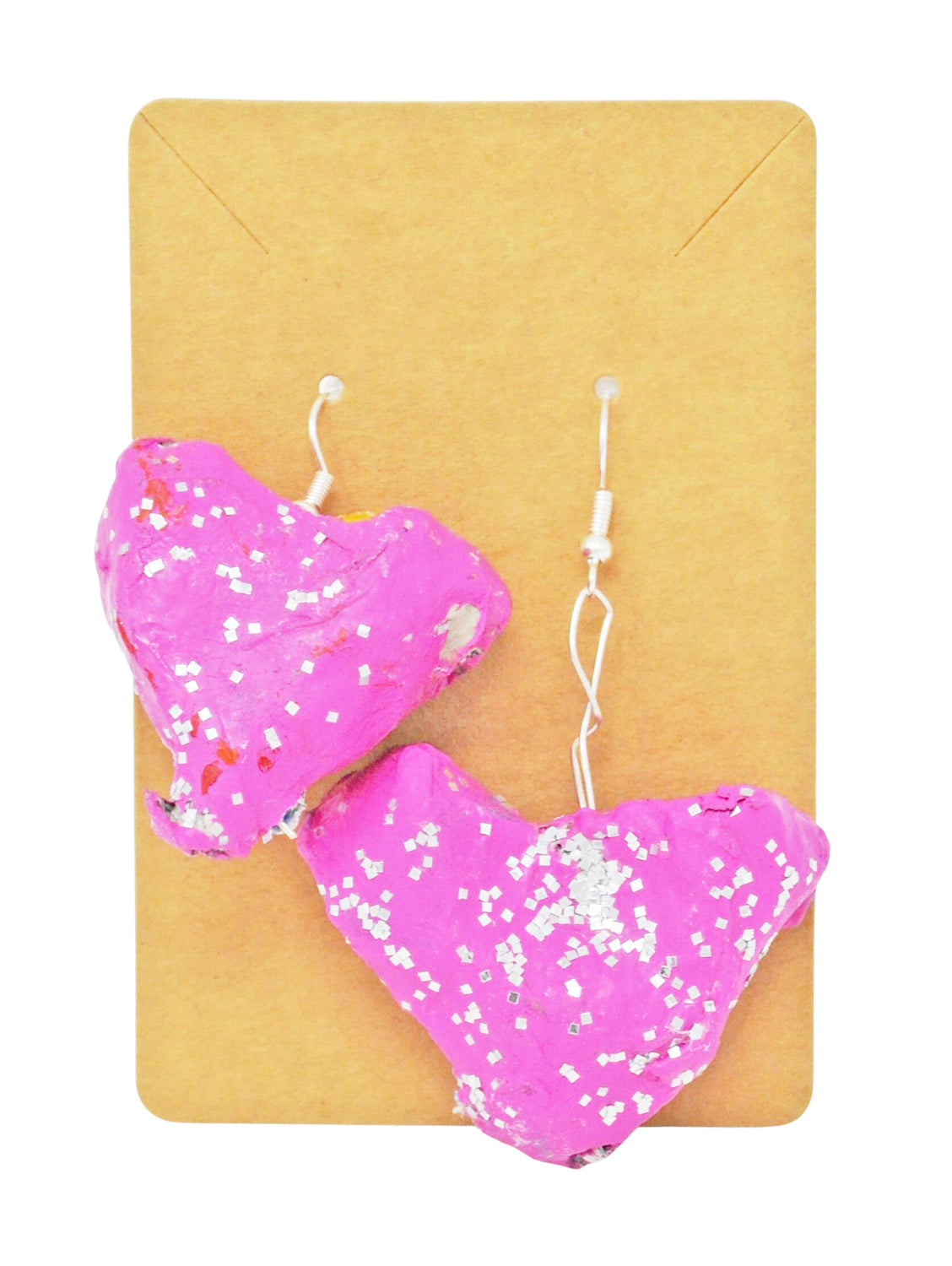 A pair of papier-mache earrings hanging from on a tan card, photographed against a white background. The earrings are 3-dimensional hearts, painted bright pink with silver glitter sprinkled across their surfaces. They have thin silver hooks.