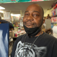 A close-up portrait photograph of a person in an art studio. The person has brown skin, short black hair, and a short black and grey beard. They are wearing a black mask, pulled down on their chin, and a black teeshirt with a white tiger head printed on the front. They look directly into the camera. A patterned banner hangs above their head. Behind them is a blurry space filled with work tables and shelves. 