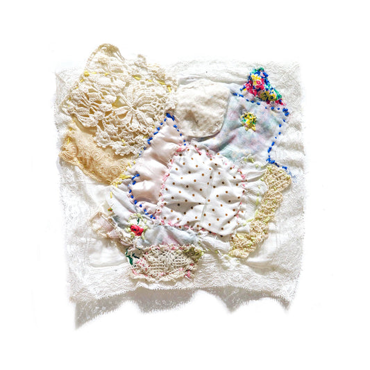 A small textile collage photographed hanging on a white wall. The base layer is a square white lace handkerchief. Pieces of aged lace and fabric swatches with polkadot or floral patterns have been stitched to the square with blue or yellow embroidery thread. 