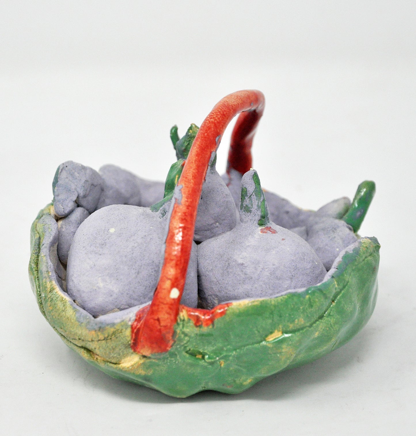 the same sculpture rotated to the right: a miniature ceramic sculpture titled Cherry Basket. The form is a shallow green bowl with a red handle arching over the top. in the bowl are grey orbs with green stems.