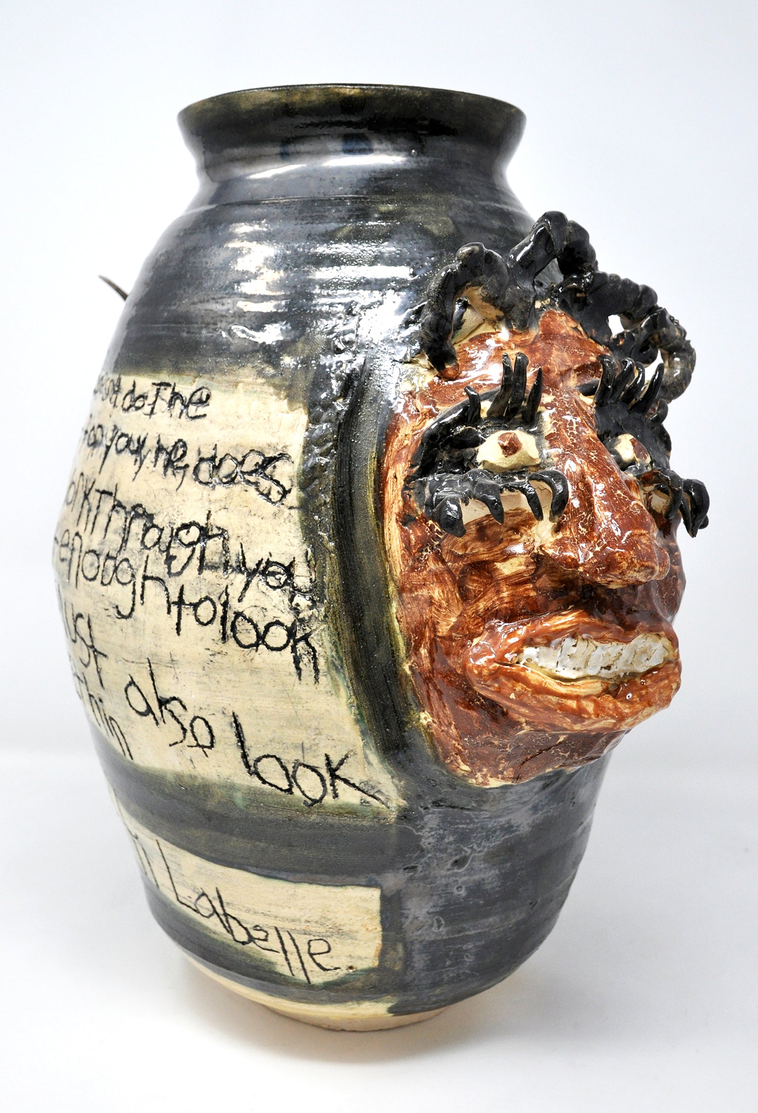 A tall ceramic vase featuring a sculpted portrait and handwritten text etched into the surface. The portrait is a smiling face painted brown with long curled eyelashes and loops of braided black hair. The portrait represents Patti Labelle. The artist has etched a long quote from the musician onto the side of the vase. Except for the head and the space around the quote, the entire vase is painted in a metallic black glaze. 