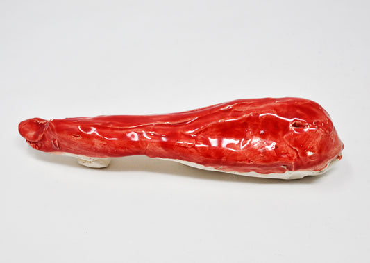 A red ceramic whale whose form tapers from the head on the right to the tail at the left of the frame. White clay can be seen at the edge along the bottom of the sculpture. 