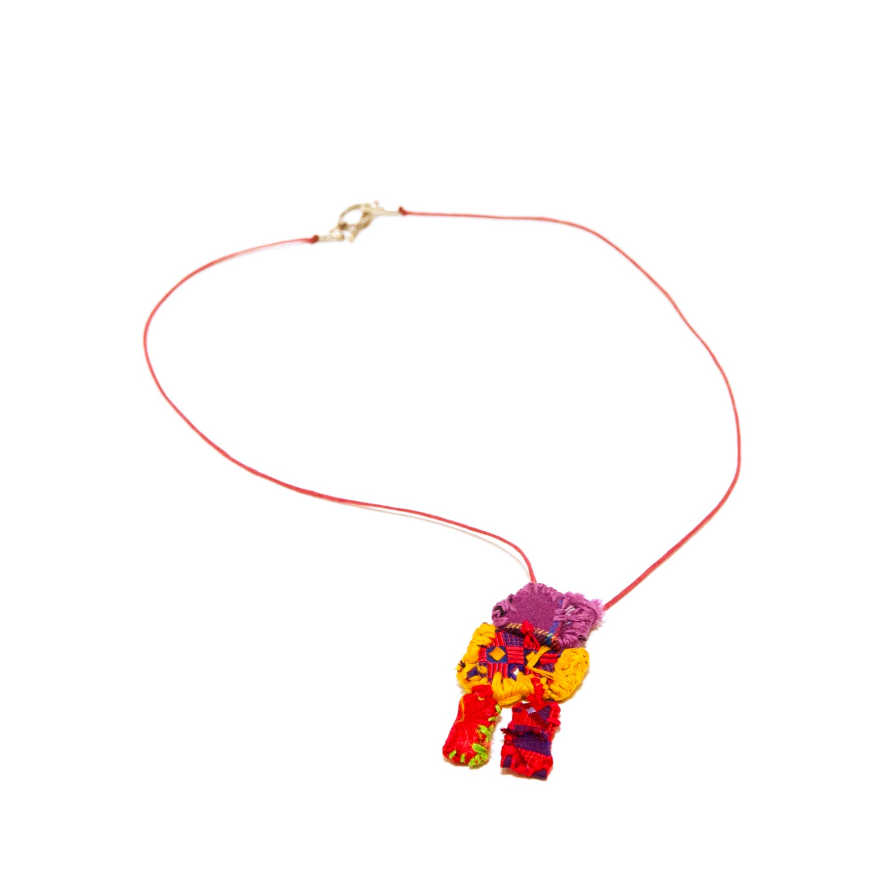 A necklace made from thread and bits of cloth, photographed on a white backdrop. A sewn object hangs from a red leather cord with a gold clasp. The object is composed of bits of red and purple cloth embroidered with yellow, green, and purple thread. The shape is vaguely figurative: a small purple square stitched to a larger red square, which is stitched to two longer rectangular limbs.