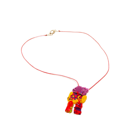 A necklace made from thread and bits of cloth, photographed on a white backdrop. A sewn object hangs from a red leather cord with a gold clasp. The object is composed of bits of red and purple cloth embroidered with yellow, green, and purple thread. The shape is vaguely figurative: a small purple square stitched to a larger red square, which is stitched to two longer rectangular limbs.