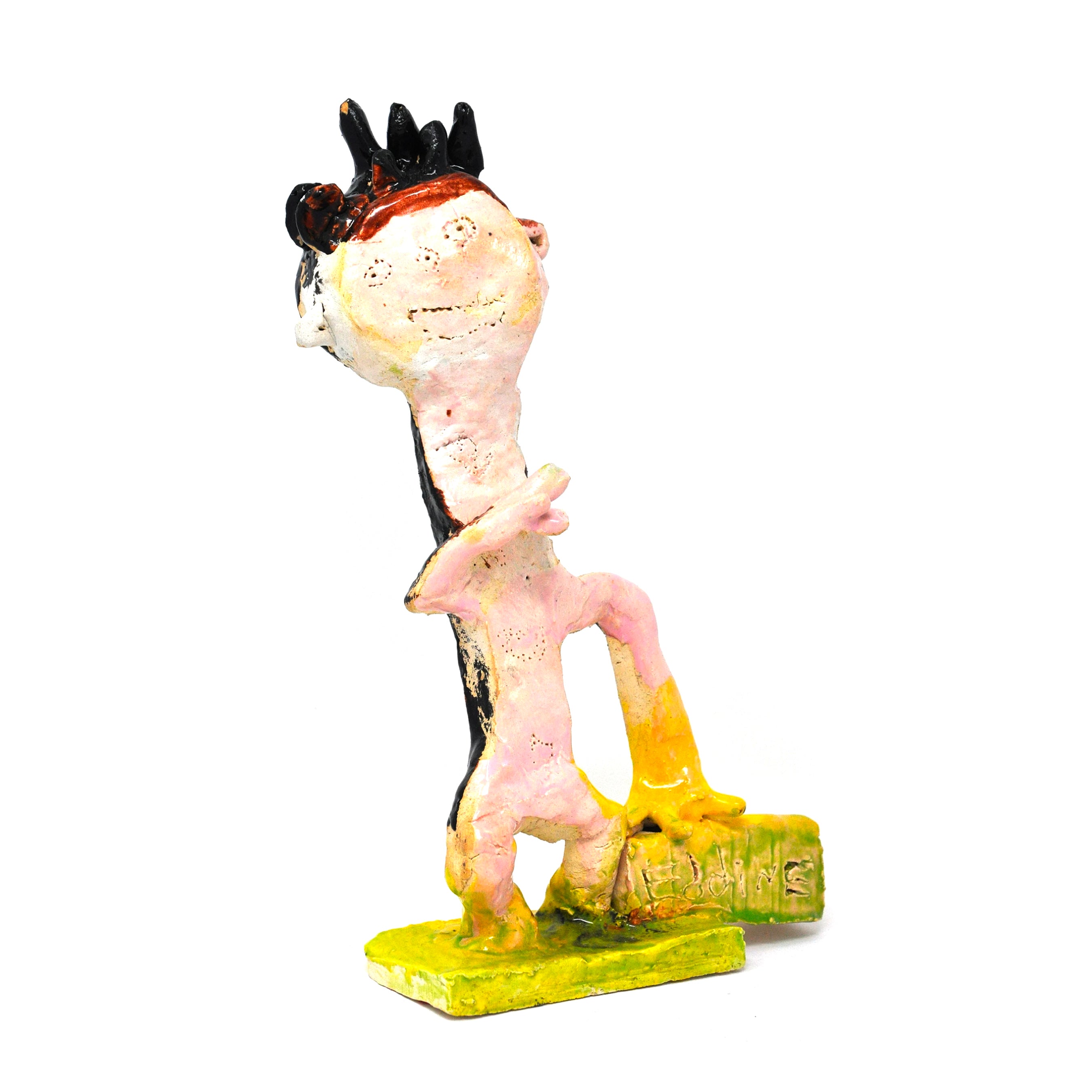 a small figurative ceramic sculpture based on the WWE wrestler Eddie Guerrero. The figure stands on a green square platform resting a hand on a plaque that says "Eddie." The figure is stylized to have a large round head and a thin rectangular body. It has spikes of hair, a smiling face etched into its head, and one arm raised by its neck. The sculpture is painted with pink and black glaze.