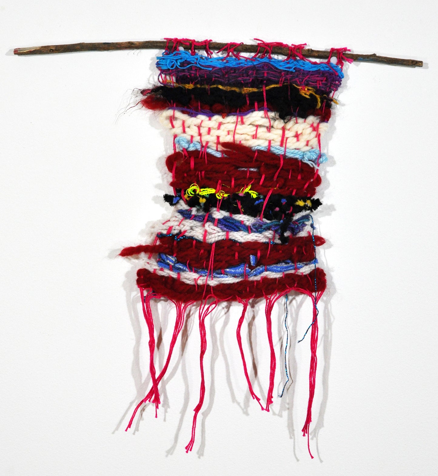 a small handwoven textile made with red and white and black yarn, hanging from a branch. Long pink tassels hang down from the bottom hem, and peak through as vertical stripes from the warp throughout the piece.