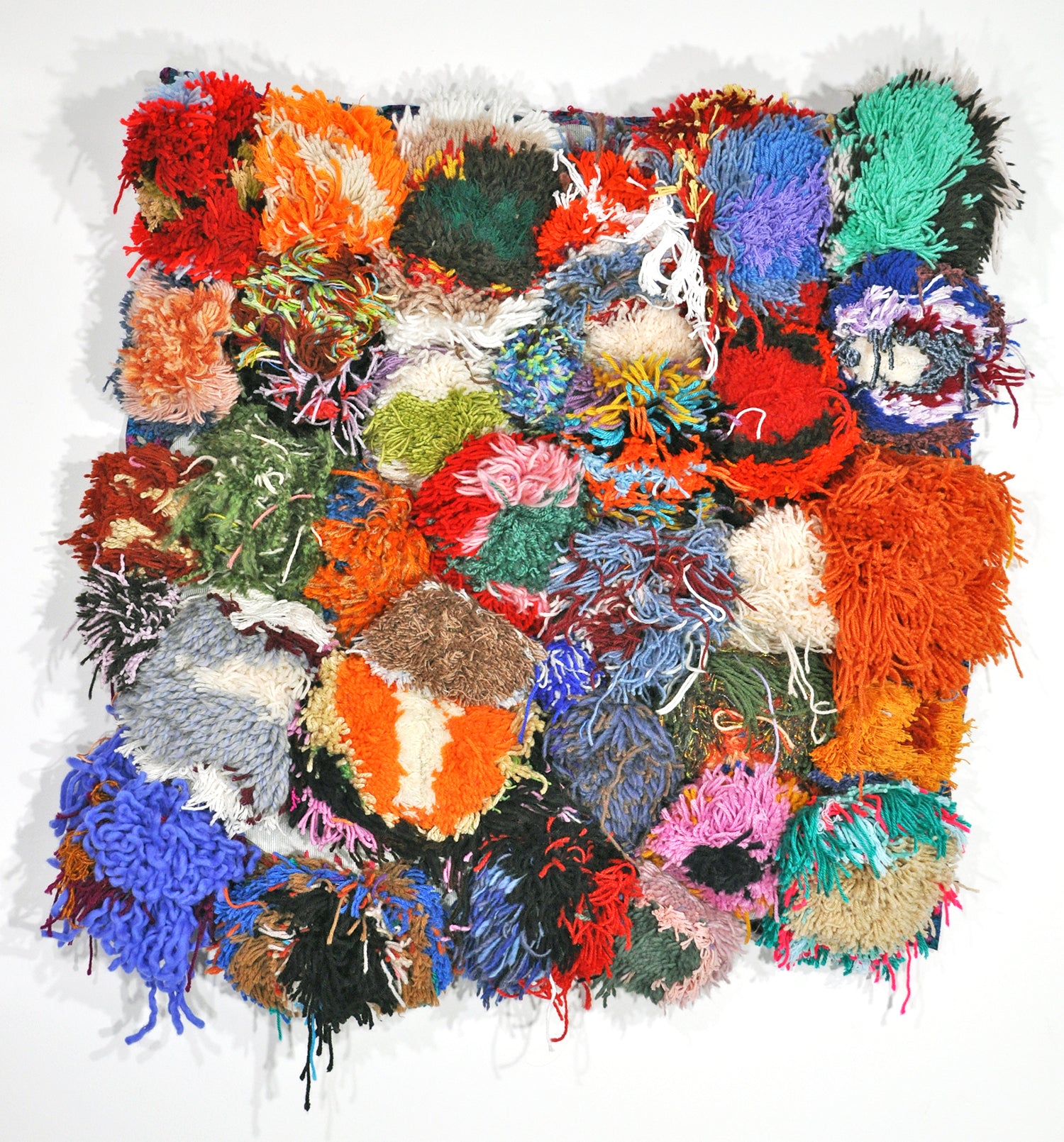 A large square fiber work photographed o a white wall. Many colorful pompoms have been sewn to a piece of fabric, covering the surface completely like a shag rug with a spotted pattern. The pompoms are variously colored orange, red, blue, pink, white, and green. Some pompoms are one solid color while others include rings of yarn in different colors and thicknesses. 