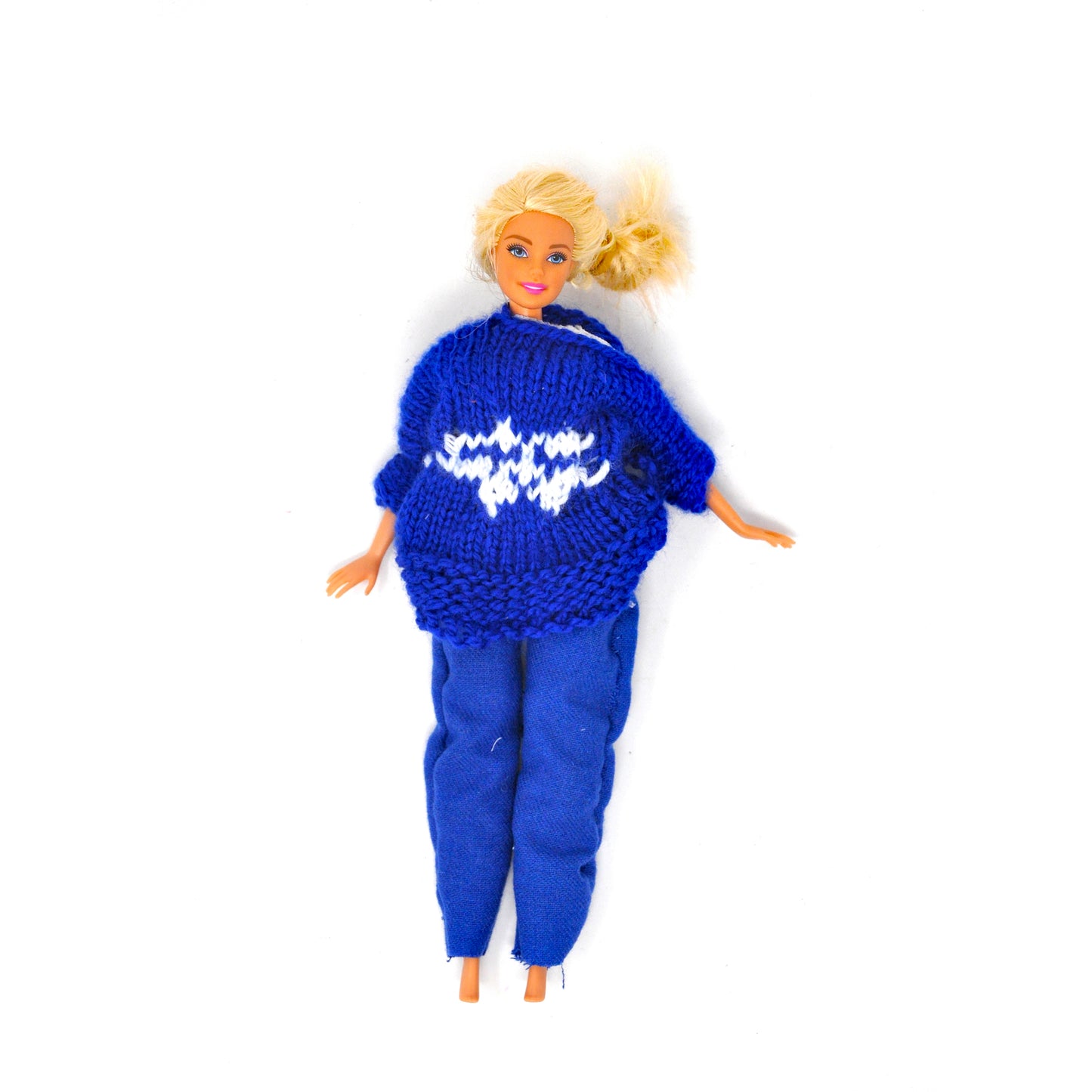 A plastic barbie doll wearing a unique handmade outfit, photographed against a white background. The barbie wears a miniature hand-knit blue fair isle sweater with a white snowflake motif in the center. The doll is also wearing hand sewn blue pants. The doll is barefoot and has blond hair and orange skin.