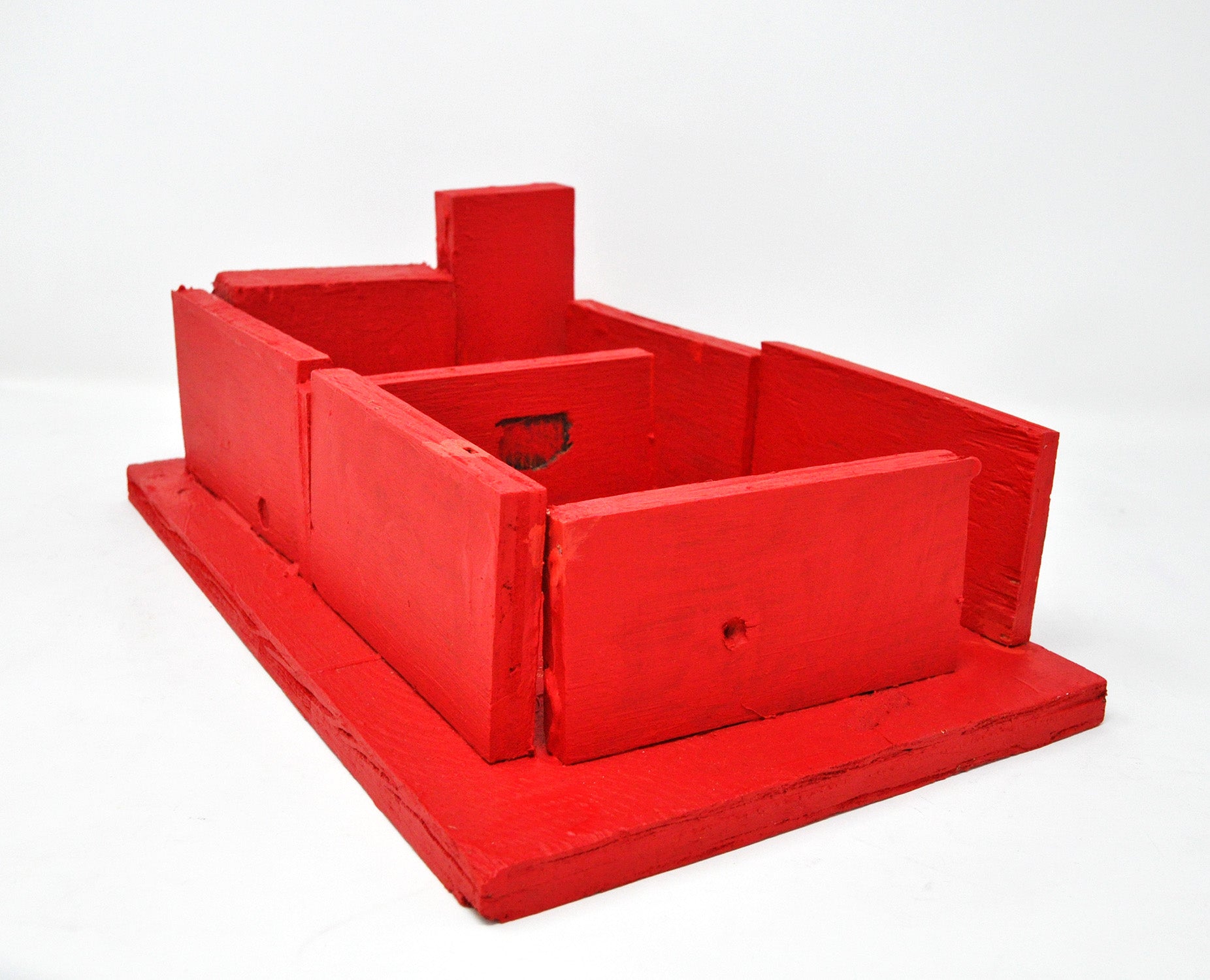 a sculpture made from wooden blocks arranged in a sort of barricade and adhered to a flat wooden plank. The structure represents a building floor plan, with no roof so the interior is visible from above. The entire wooden structure has been painted bright red. This work is part of a larger series of miniature buildings and city scapes that the artist often activates with small figurines.