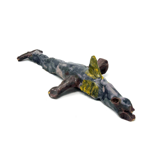 A small ceramic sculpture of a sea creature, photographed on a white background. The creature resembles a dolphin or a shark. It has side and tail fins, an open mouth, and a long tubular body coated in a purple and pink glaze. It has gils etched onto its side and a pointed yellow fin on its back.