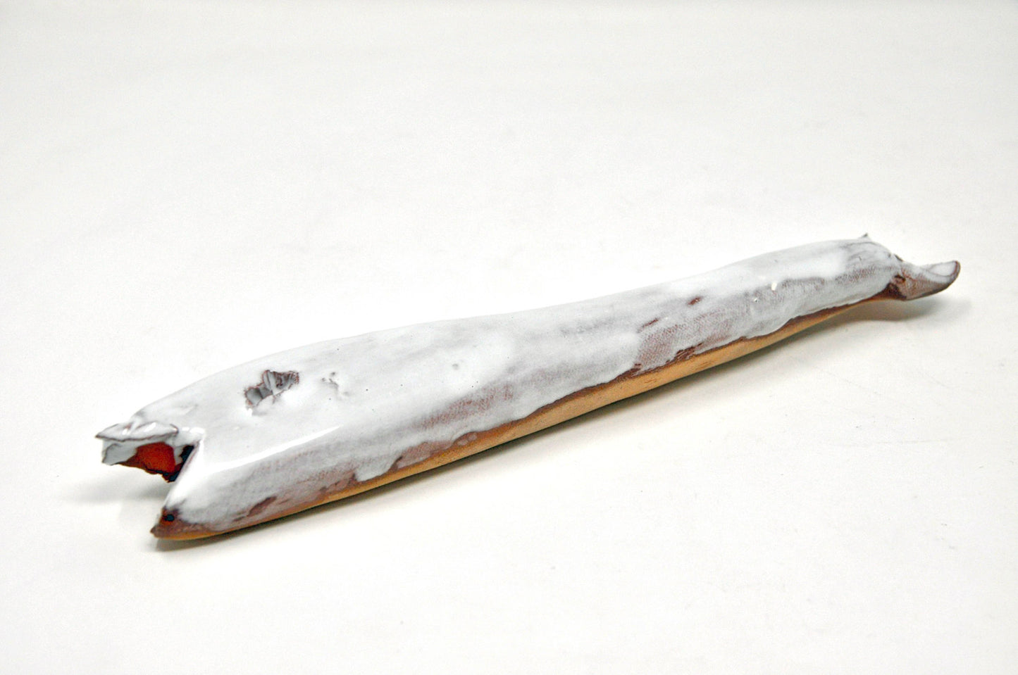 A long white ceramic whale that is fairly flat. It's mouth is open, revealing a hint of red glaze, and the natural clay body is visible along the bottom edge of the whale that rests on the ground. 