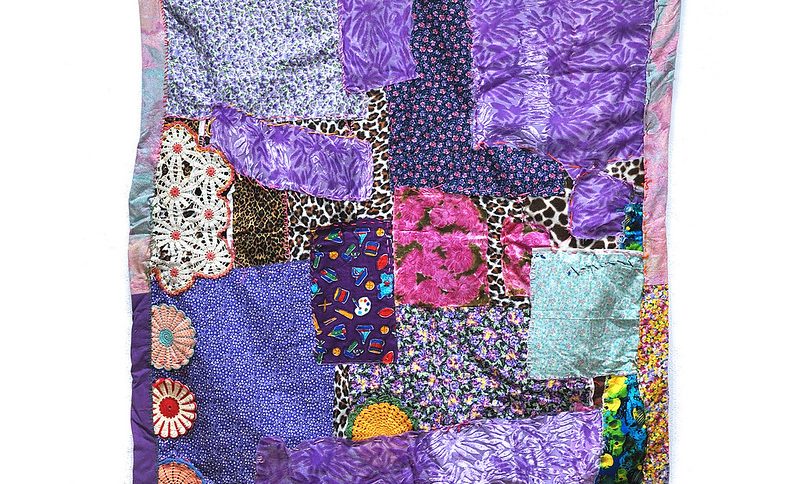 A collage of textiles of varying square and rectangle sizes, patterns and colors, layered on top of one another with some round, multi-colored doilie haped patches and a patchwork border.