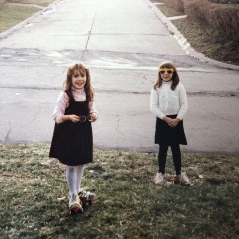 A photograph of two young girls, one wearing a black pinafore, the other yellow-rimmed sunglasses, standing on a patch of grass at the end of a suburban street.