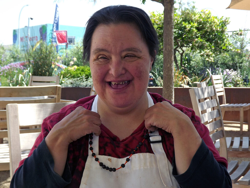 A portrait of a woman wearing a red shirt and white apron showing the viewer her necklace