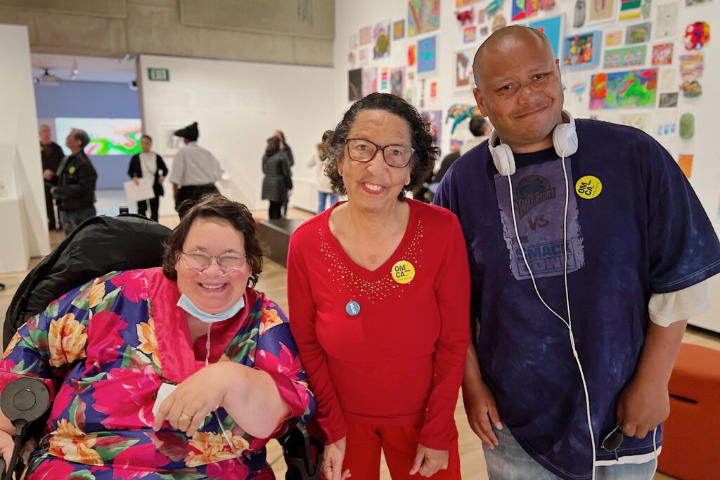 Three NIAD artists smile at the camera at a gallery opening. Rebecca Jantzen is seated in a wheelchair wearing a bright floral dress. Dorian Reid stands in the center with glasses, a bright red top and matching red lipstick, Jason Powell-Smith is to the right. Jason has white headphones around this neck and a purple t-shirt. Donate now.