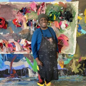 NIAD artist Peter Harris smiles for the camera wearing a beanie, black apron and standing in front of his fiber art. Donate to NIOAD today.
