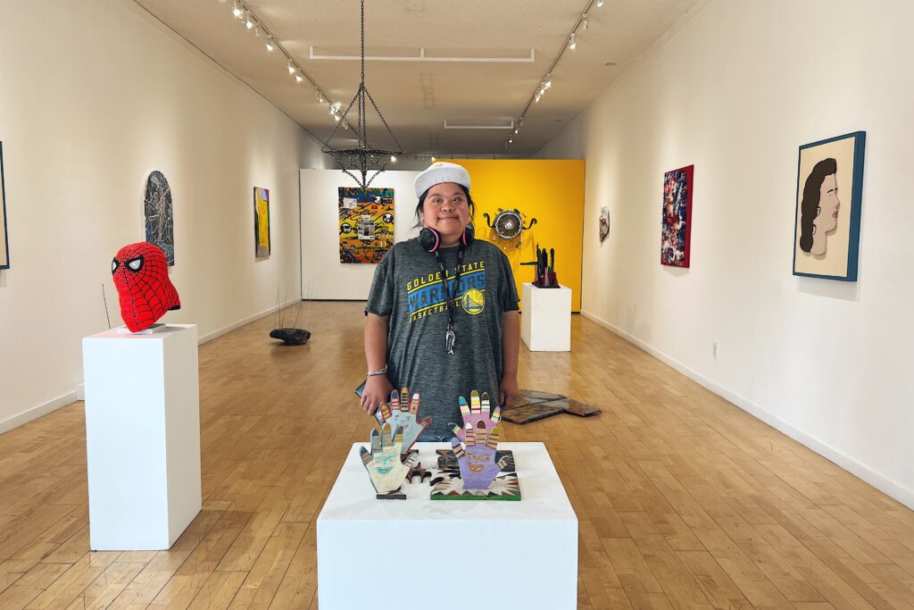 A dark haired woman with a white baseball cap, grey t-shirt with yellow and blue lettering stands at the front of a large art gallery, filled with all different sizes and kinds of art. Including a bust of Spiderman, a sculpture of different pastel hands, and in the back a yellow wall with a sculpture make of a wheel.