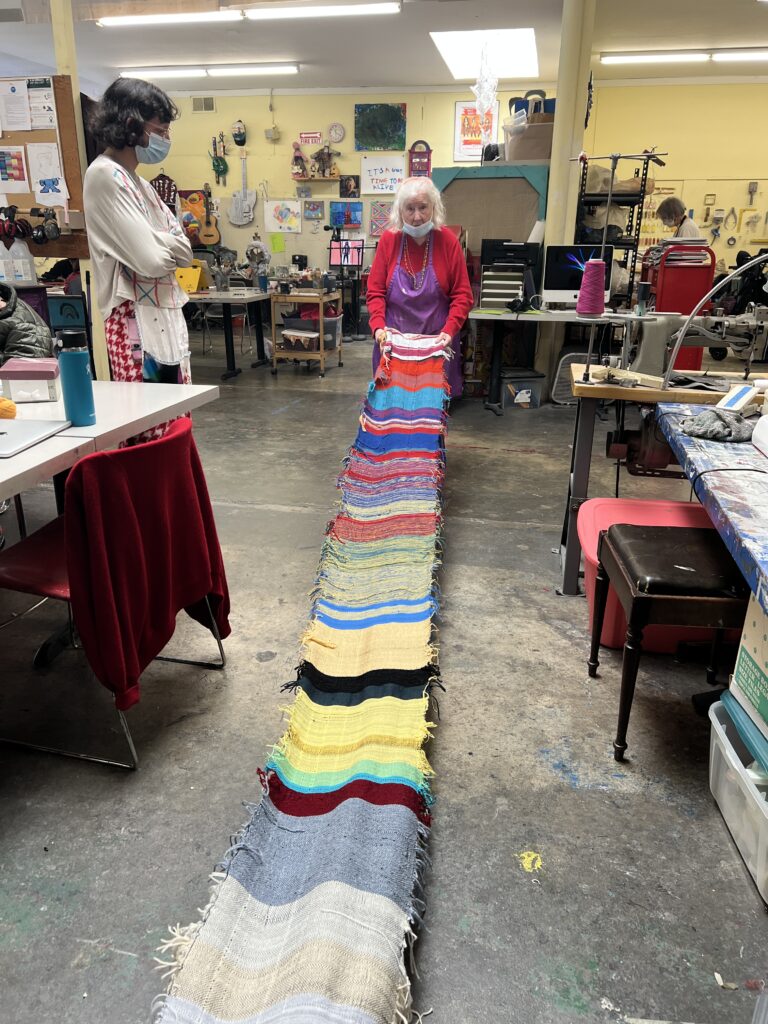 a woman with grey hair, red sweater and a purple apron holds one side of a very long multi-colored weaving that has strips of different colors and widths of yarn. The other side of the weaving comes towards the viewer and ends outside of the frame. There are many ways to give to NIAD Art Center.