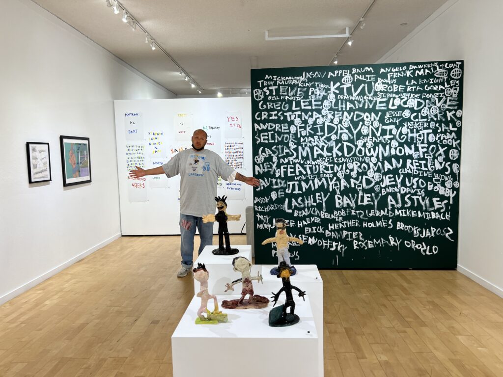 A tall, blad man with a mask under his chin, wearing a long grey t-shirt, jeans and trainers, stands in the middle of a gallery art show with his arms outstretched. In front of him are sculptures of people, to the side of him is a wall sized dark green mural with white lettering all over it.
