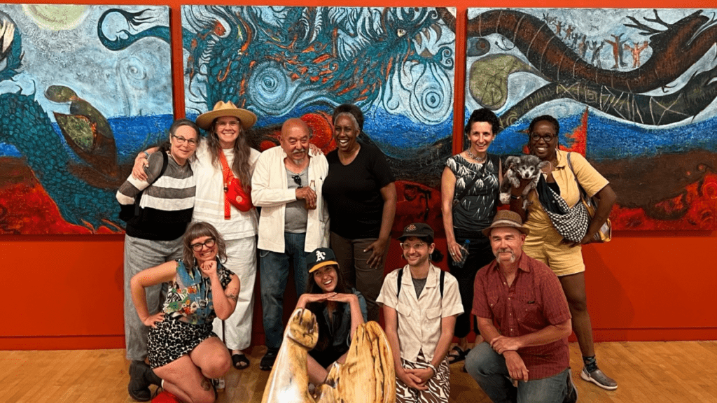 Two rows of smiling NIAD staff past and present pose for a group photo in front of an epic triptych depicting large dragons and small humans, behind a smooth, shiny wool sculpture, both works by NIAD facilitator Andrés Cisneros-Galindo at his Richmond Art Center exhibition "Nahui Ollin". Large sans serif yellow text superimposed over the picture reads "NIAD IS HIRING".