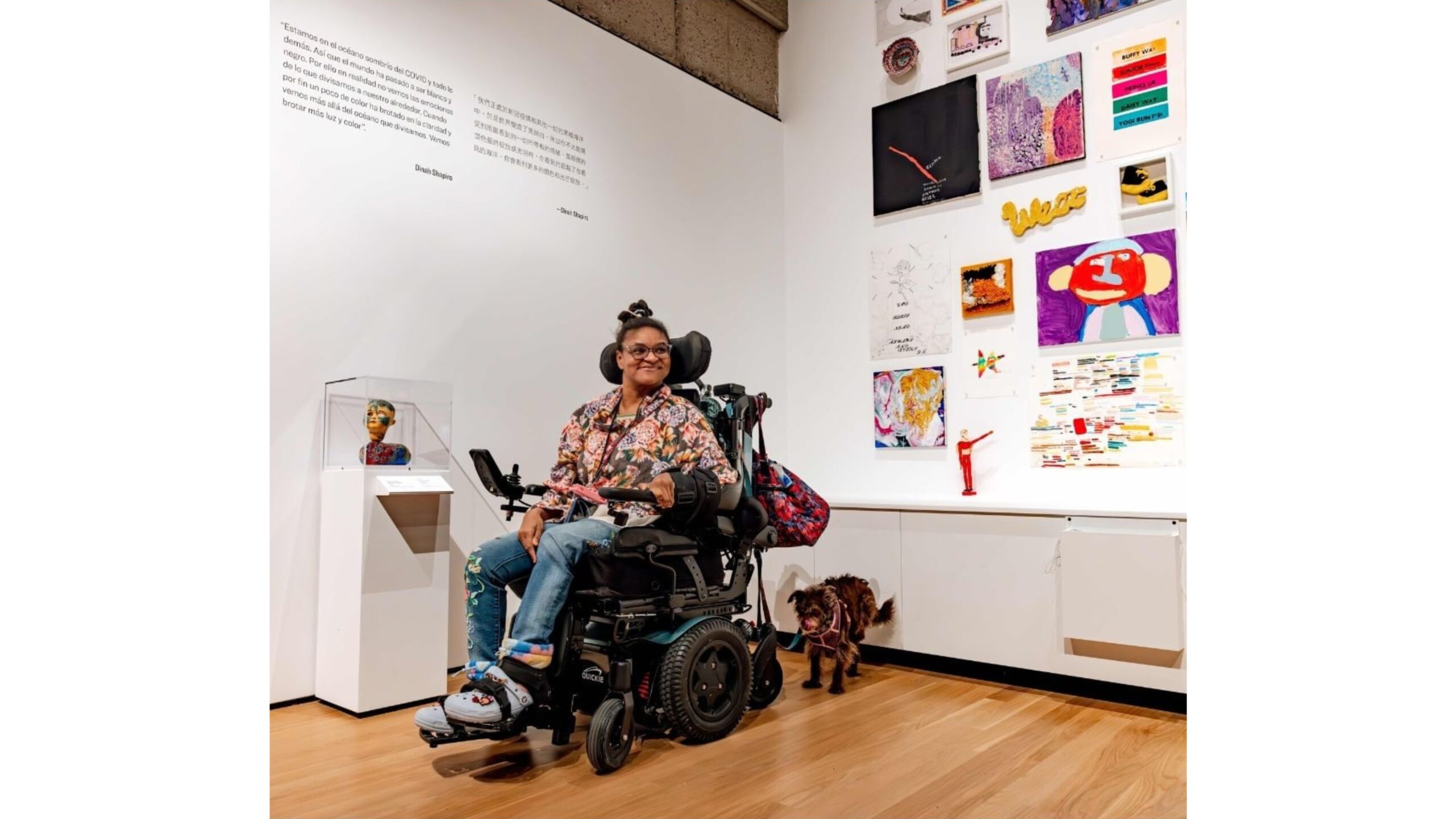 an artist with dark skin in a wheelchair and a companion dog smiles in a gallery setting