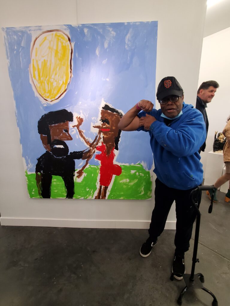 NIAD artist Arstanda Billy White - a man with dark brown skin - wearing an SF giants hat, showing his bicep in front of one of his paintings depicting two figures in a landscape, in an art fair setting.