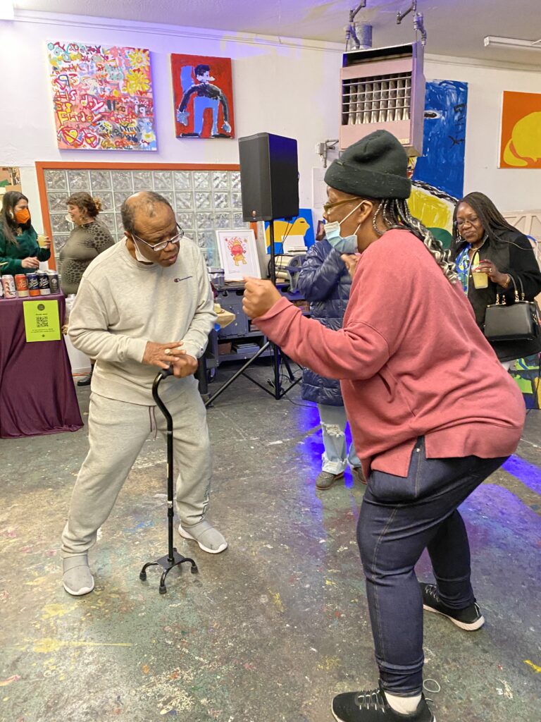 NIAD artist Arstanda Billy White - a man with dark brown skin wearing a gray sweatsuit holding a cane - dances with NIAD artist Felicia Griffin - a woman with dark brown skin wearing a black beanie - at NIAD's Win Win 11 party.