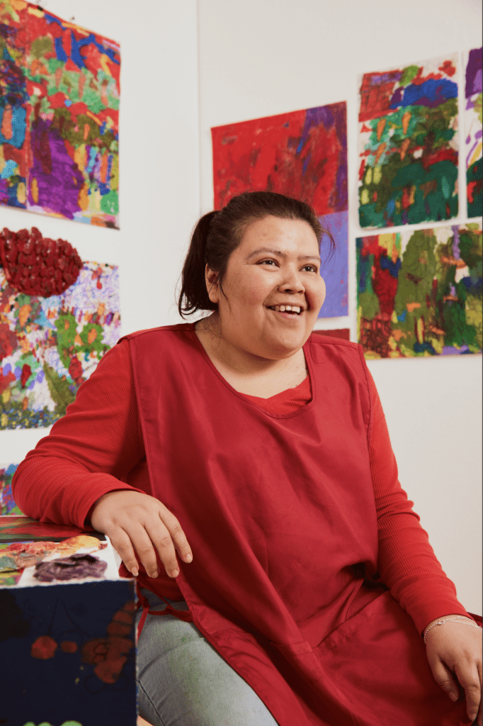 NIAD artist Karla Alfaro - a woman with light brown skin wearing a loose red tunic - smiles and looks off camera as she poses in front of a wall of her artwork.