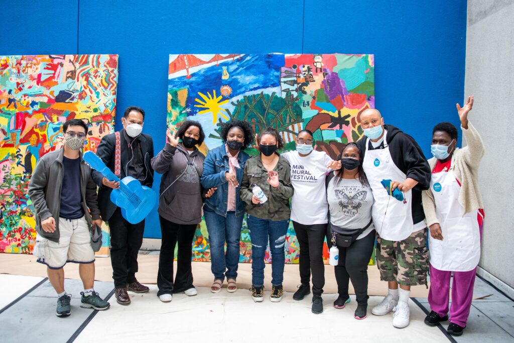 a group of NIAD artists standing in front of a colorful mural against a bright blue background at the SFMOMA mini mural festival.